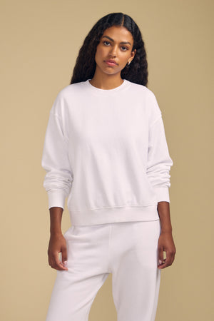 A woman stands against a beige background, wearing the YOKO SWEATSHIRT by Velvet by Graham & Spencer in plain white cotton fleece paired with matching white pants, looking directly at the camera with a neutral expression. The relaxed silhouette of her outfit enhances its understated appeal.