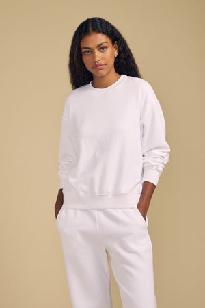 A person with long curly hair stands against a beige background, wearing a plain white YOKO SWEATSHIRT by Velvet by Graham & Spencer and matching white sweatpants with hands in the pockets, showcasing a relaxed silhouette in soft cotton fleece.