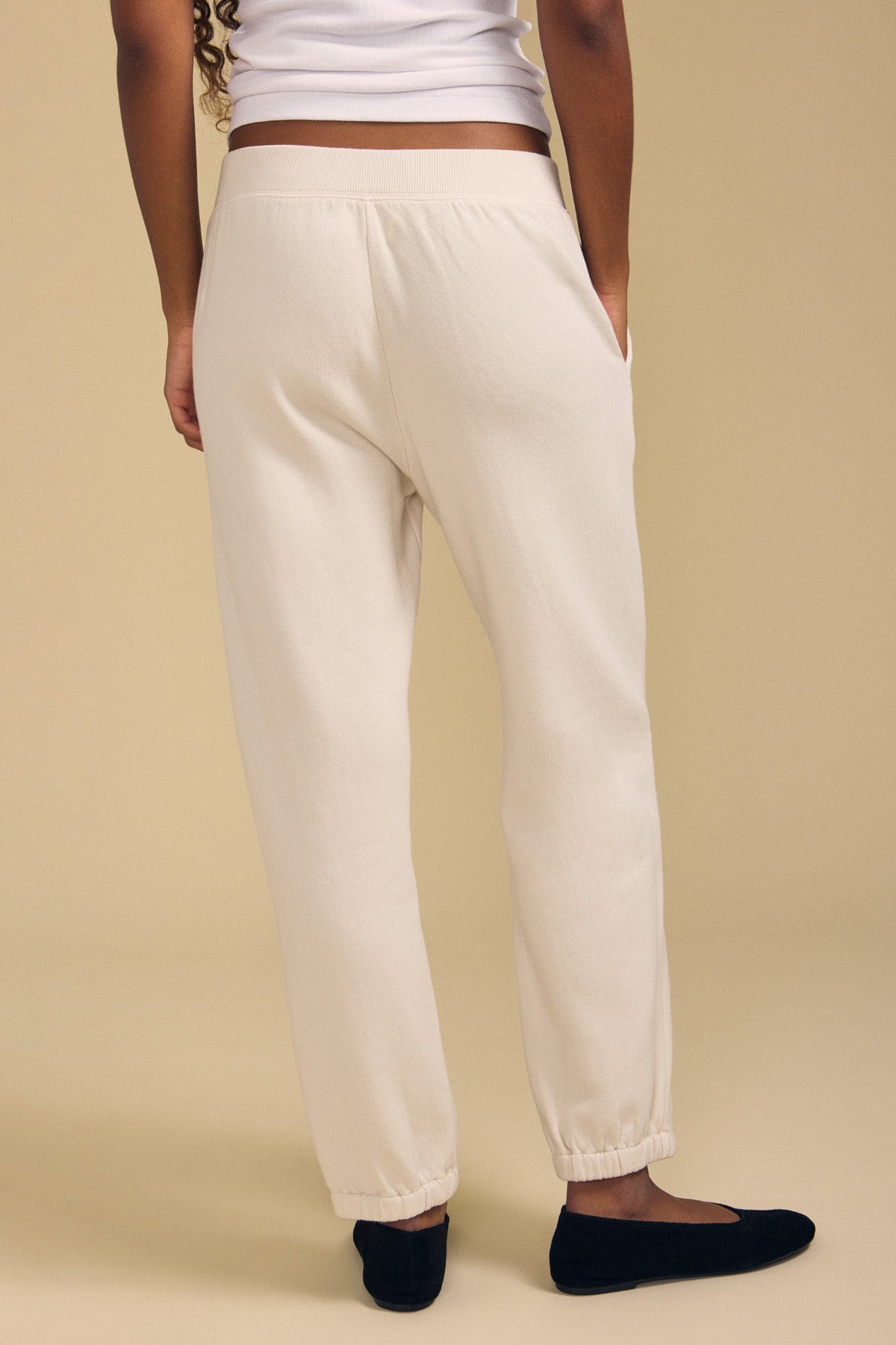 Wearing the ZARA SWEATPANT by Velvet by Graham & Spencer—off-white jogger-style sweatpants in a relaxed fit cotton fleece—a person stands against a beige background, shown from the back, paired with a white top and black flat shoes.