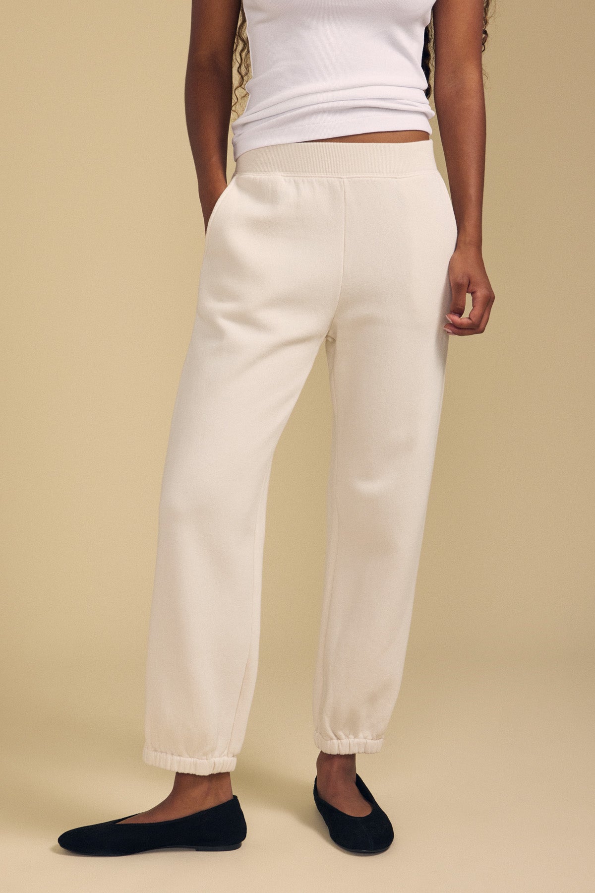 Wearing the ZARA SWEATPANT joggers by Velvet by Graham & Spencer in a cream hue and relaxed fit, paired with a white sleeveless top and black flats, this look stands out against a beige background. The cotton fleece fabric of the sweatpants delivers both comfort and style to the ensemble.