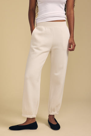 Wearing the ZARA SWEATPANT joggers by Velvet by Graham & Spencer in a cream hue and relaxed fit, paired with a white sleeveless top and black flats, this look stands out against a beige background. The cotton fleece fabric of the sweatpants delivers both comfort and style to the ensemble.