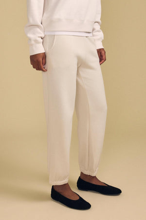 A person wearing the ZARA SWEATPANT by Velvet by Graham & Spencer in off-white cotton fleece, paired with a matching sweatshirt and black ballet flats, standing on a beige background.