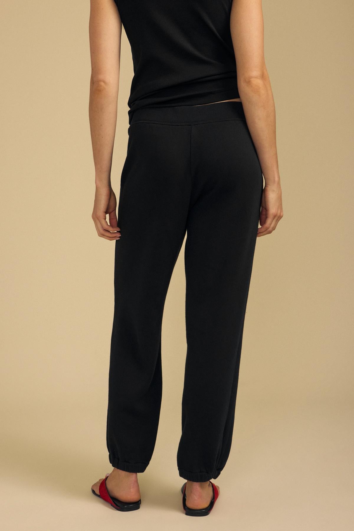 A person wearing a black short-sleeve shirt, Velvet by Graham & Spencer ZARA SWEATPANT in black, and red sandals stands facing away on a beige background.-41980701966529