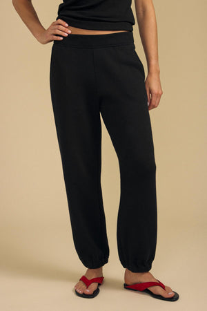 A person stands against a beige background, showing only the lower half of their body while wearing relaxed fit black ZARA SWEATPANT by Velvet by Graham & Spencer, paired with a black shirt and red sandals. Crafted from soft cotton fleece, these sweatpants provide both comfort and style.