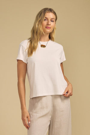 A woman stands against a plain beige background, her right hand holding the hem of her shirt, wearing Velvet by Graham & Spencer’s TIA TEE—a timeless white cotton jersey crewneck—paired with beige pants.