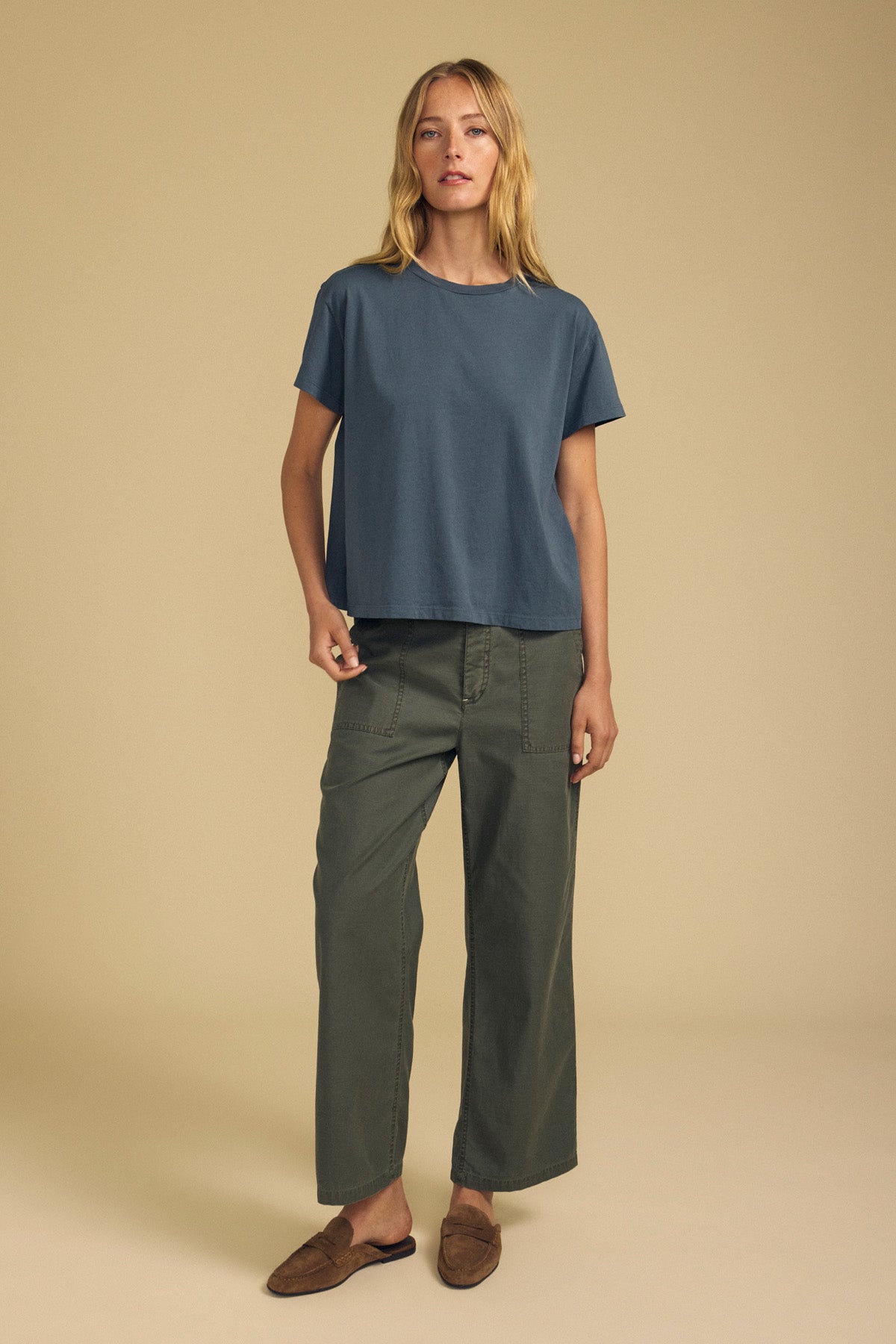 A woman stands against a beige background wearing the TIA TEE by Velvet by Graham & Spencer—a loose blue cotton jersey crewneck tee, effortlessly timeless—paired with olive green wide-leg pants and brown slip-on shoes.-41964724682945