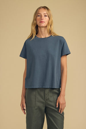 A woman stands against a beige background, wearing the TIA TEE by Velvet by Graham & Spencer—an effortlessly timeless crewneck crafted from soft blue cotton jersey—paired with olive green pants, looking straight at the camera with a neutral expression.