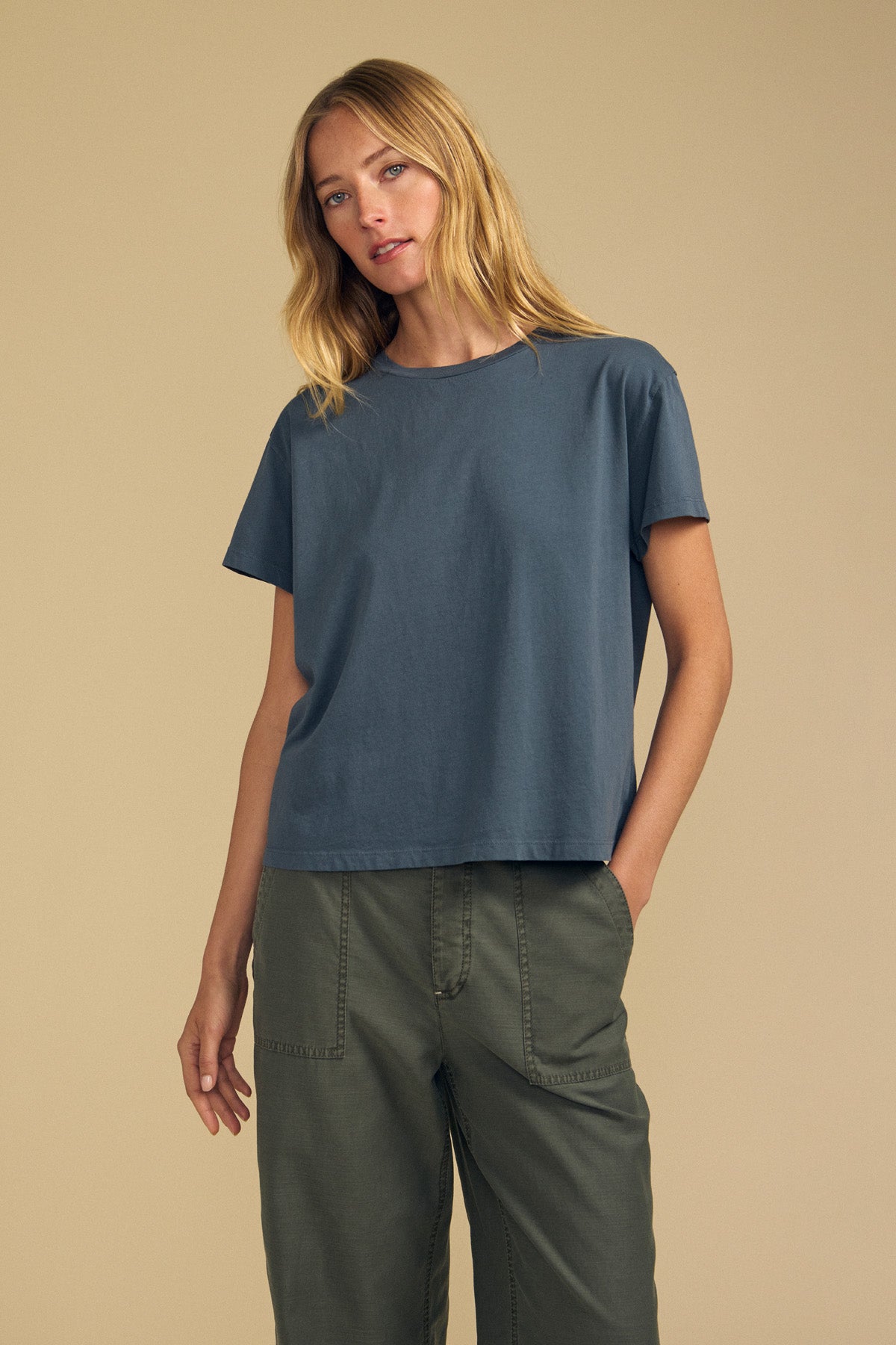 Model wears the TIA TEE by Velvet by Graham & Spencer—a loose-fitting blue crewneck top crafted from soft cotton jersey—paired with olive green pants, standing against a plain beige backdrop. This timeless essential elevates any wardrobe.
