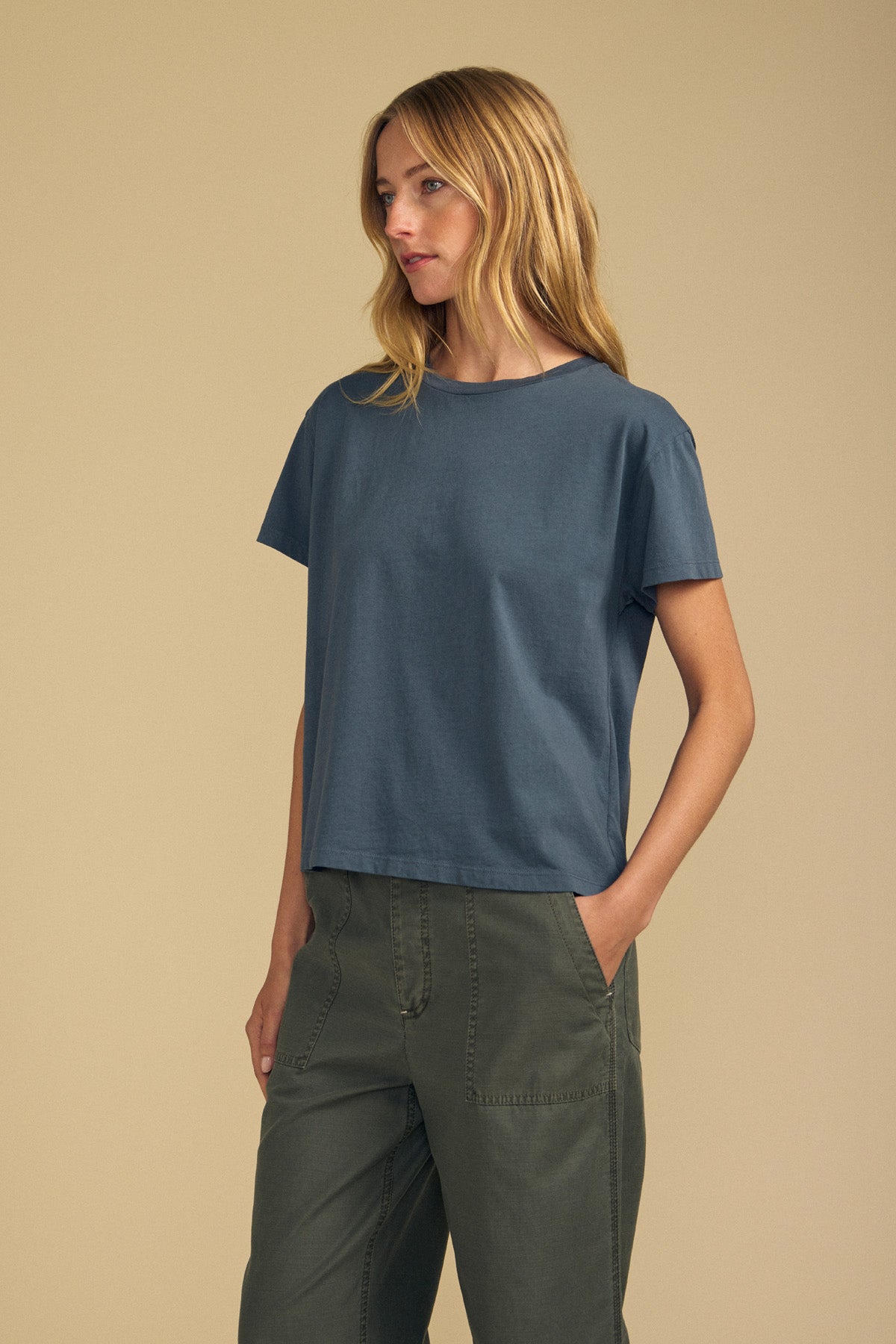A woman stands against a beige background, wearing the loose-fitting TIA TEE by Velvet by Graham & Spencer in blue cotton jersey and green cargo pants, with her hands in her pockets and looking to the side.
