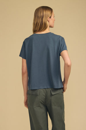 A person with long, light brown hair stands facing away, wearing the Velvet by Graham & Spencer TIA TEE—a loose blue cotton jersey crewneck—paired with olive green pants against a plain beige background. This tee is a timeless essential for effortless style.