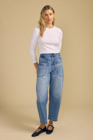 A person wearing the CAMINO TEE by Velvet by Jenny Graham—a fitted white long-sleeve modal rib shirt—paired with high-waisted blue jeans and black flats stands against a plain beige background. This versatile layering piece offers a slimmer fit for a sleek, modern silhouette.