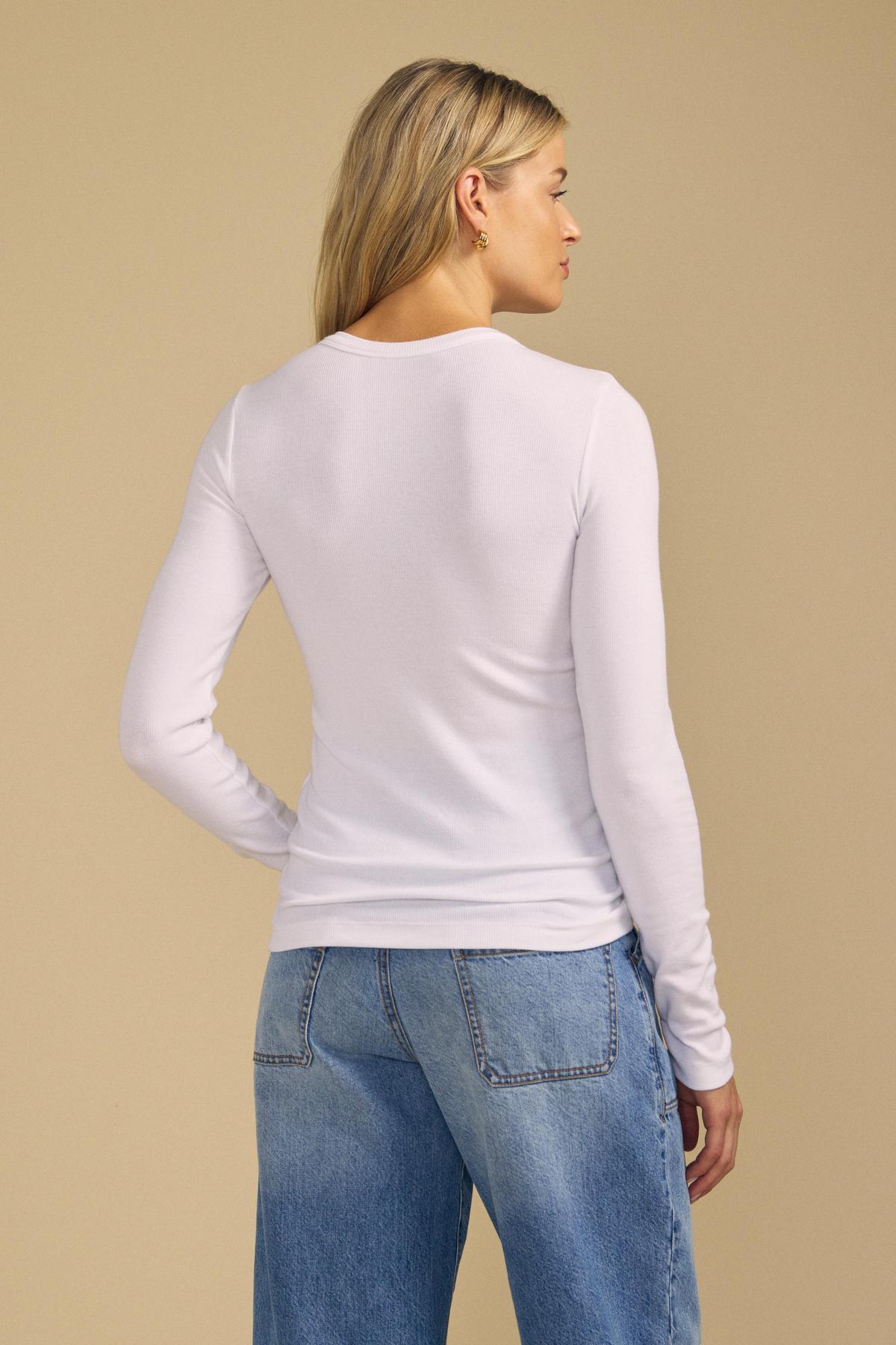 A person stands with their back to the camera, wearing the CAMINO TEE by Velvet by Jenny Graham—a long-sleeve white shirt made from soft modal rib for a slimmer fit and versatile layering—paired with blue jeans, set against a plain beige background.