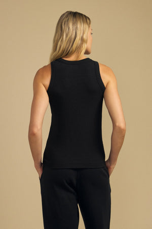 A person with long blonde hair stands with their back to the camera against a plain beige background, wearing the CRUZ TANK TOP by Velvet by Jenny Graham and black pants—a timeless look perfect for any capsule wardrobe.