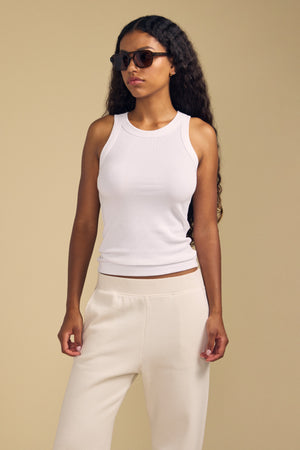 A woman with long curly hair wearing sunglasses, the Velvet by Jenny Graham CRUZ TANK TOP, and cream-colored pants stands against a plain beige background—a timeless capsule wardrobe look crafted from soft cotton-modal fabrics.