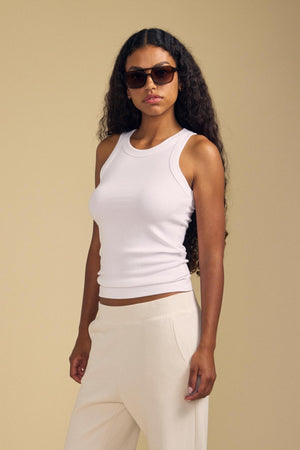 A woman with long curly hair, wearing sunglasses, cream pants, and the Velvet by Jenny Graham CRUZ TANK TOP—an essential ribbed cotton-modal piece for any capsule wardrobe—stands against a beige background.