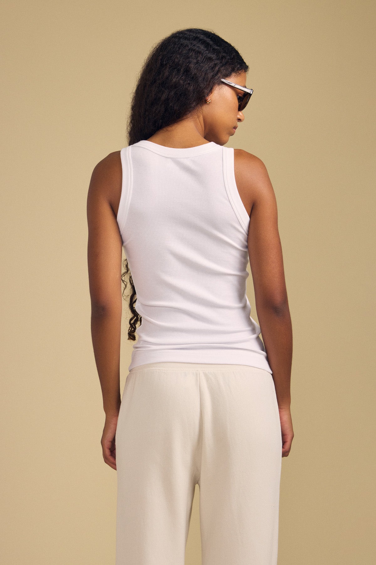 A person with long curly hair wearing sunglasses and cream pants stands with their back to the camera against a beige background, dressed in the CRUZ TANK TOP by Velvet by Jenny Graham.