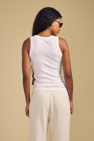 A person with long curly hair wearing sunglasses and cream pants stands with their back to the camera against a beige background, dressed in the CRUZ TANK TOP by Velvet by Jenny Graham.