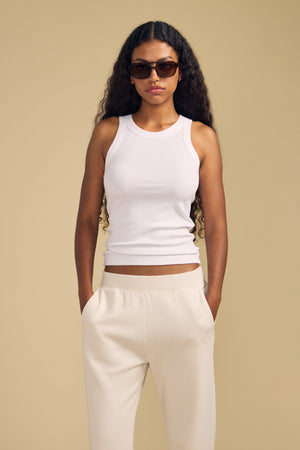 A woman wearing sunglasses, the CRUZ TANK TOP by Velvet by Jenny Graham—crafted from a soft cotton-modal blend in white—and cream-colored pants stands with her hands in her pockets against a beige background, showcasing an effortless look perfect for any capsule wardrobe.