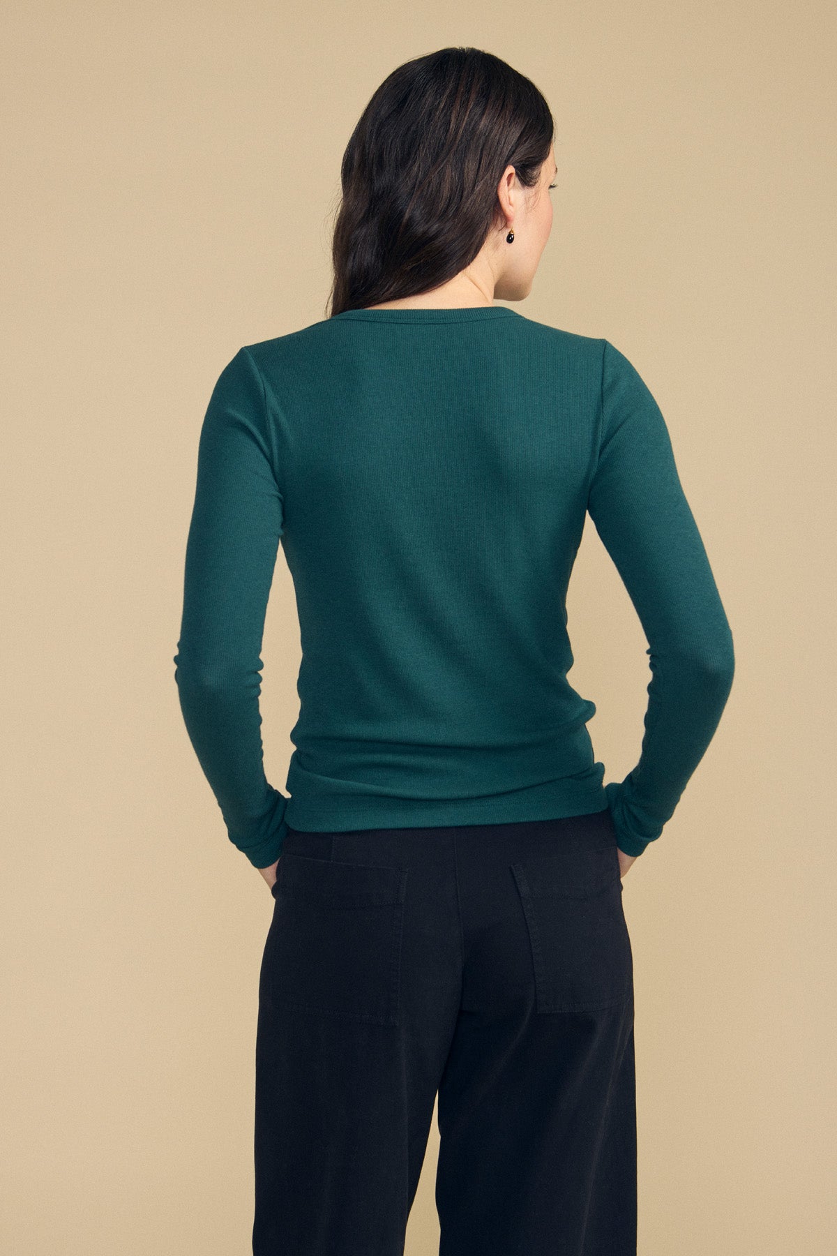 A person stands with their back to the camera, wearing the Velvet by Jenny Graham CAMINO TEE—a slim fit, long-sleeve green layering top—paired with black pants against a beige background.-41860764631233