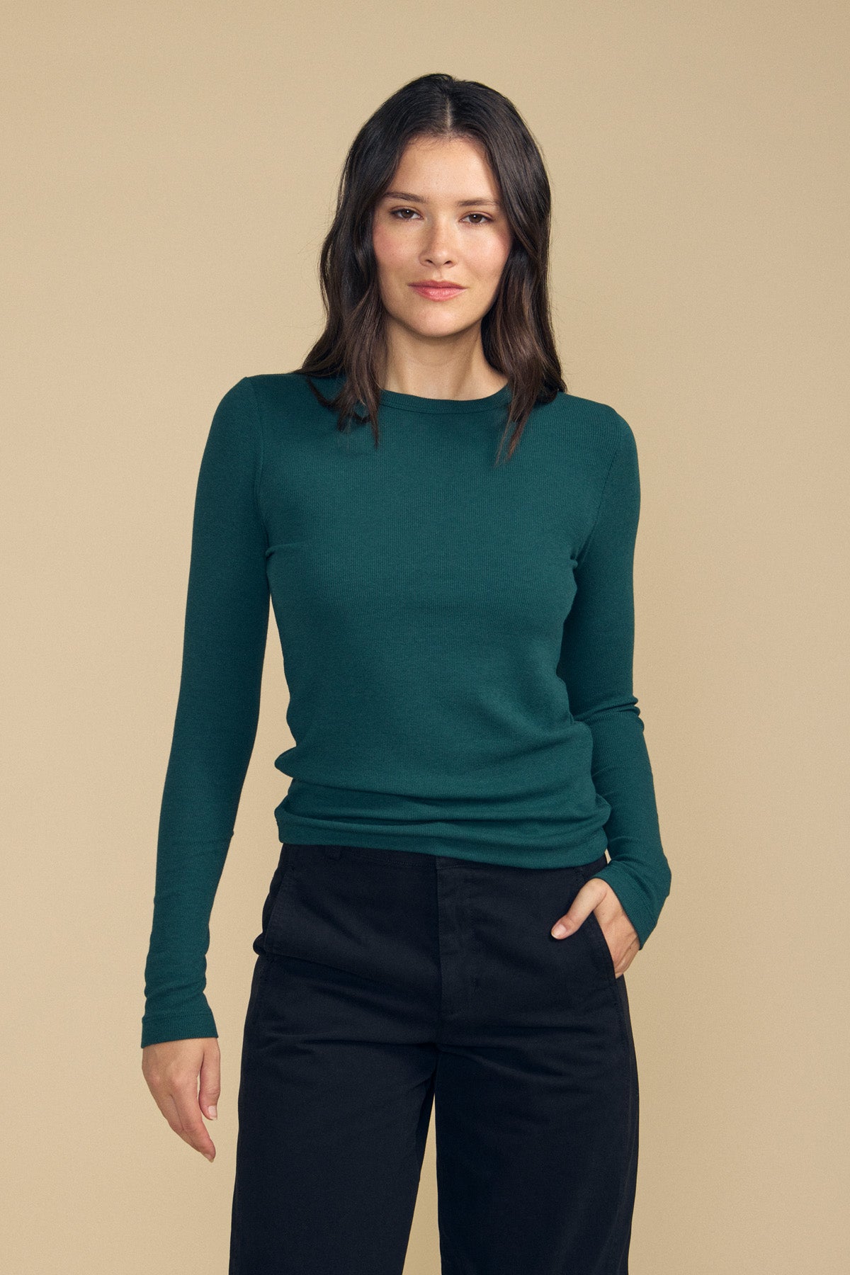 A woman with medium-length dark hair wears the Velvet by Jenny Graham CAMINO TEE—a slim fit green long-sleeve layering top—paired with black pants, standing against a plain beige background with one hand in her pocket.-41860764598465