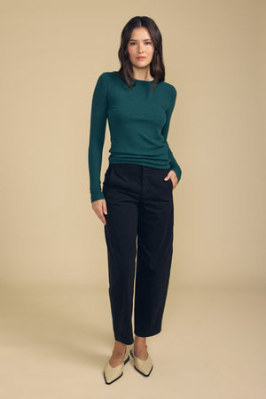 A woman stands against a beige background wearing a green long-sleeve top, the Velvet by Graham & Spencer TENLEY PANT in black high-waisted style, and cream-colored pointed shoes.