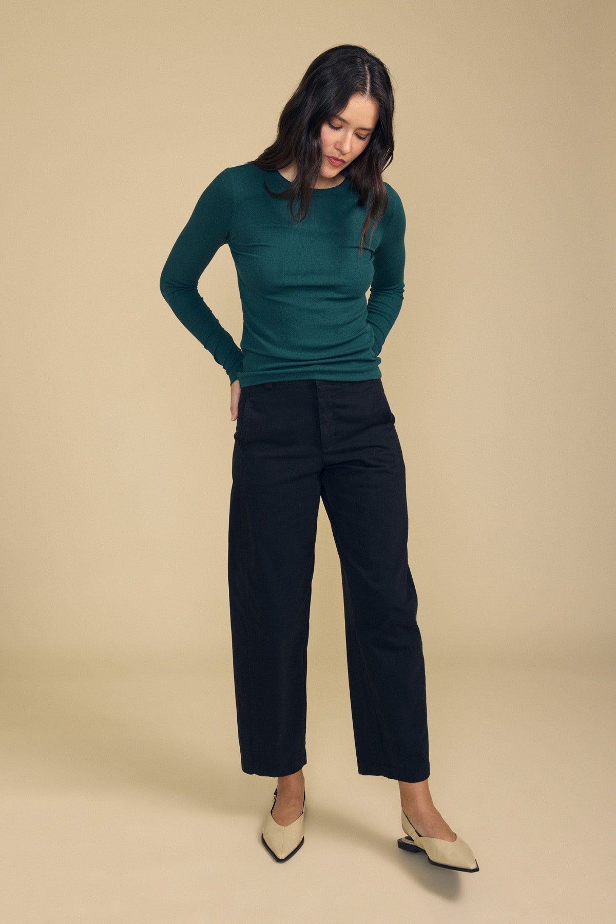 A person wearing a long-sleeve green top, the TENLEY PANT by Velvet by Graham & Spencer in black high-waisted wide-leg style, and beige shoes stands on a beige background, looking down with their hands in their pockets.