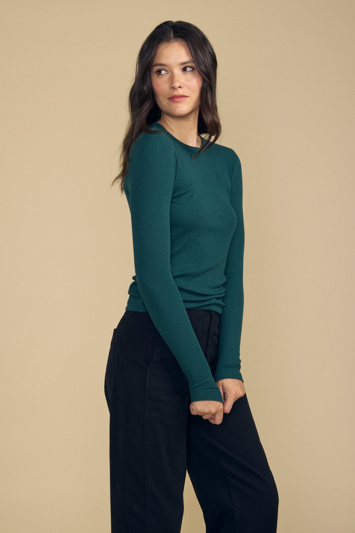 A woman with long brown hair wears the Velvet by Jenny Graham CAMINO TEE—a teal, slim fit, long-sleeve layering top crafted from soft modal rib—paired with black pants, standing against a plain beige background and looking to her left.-41860764664001