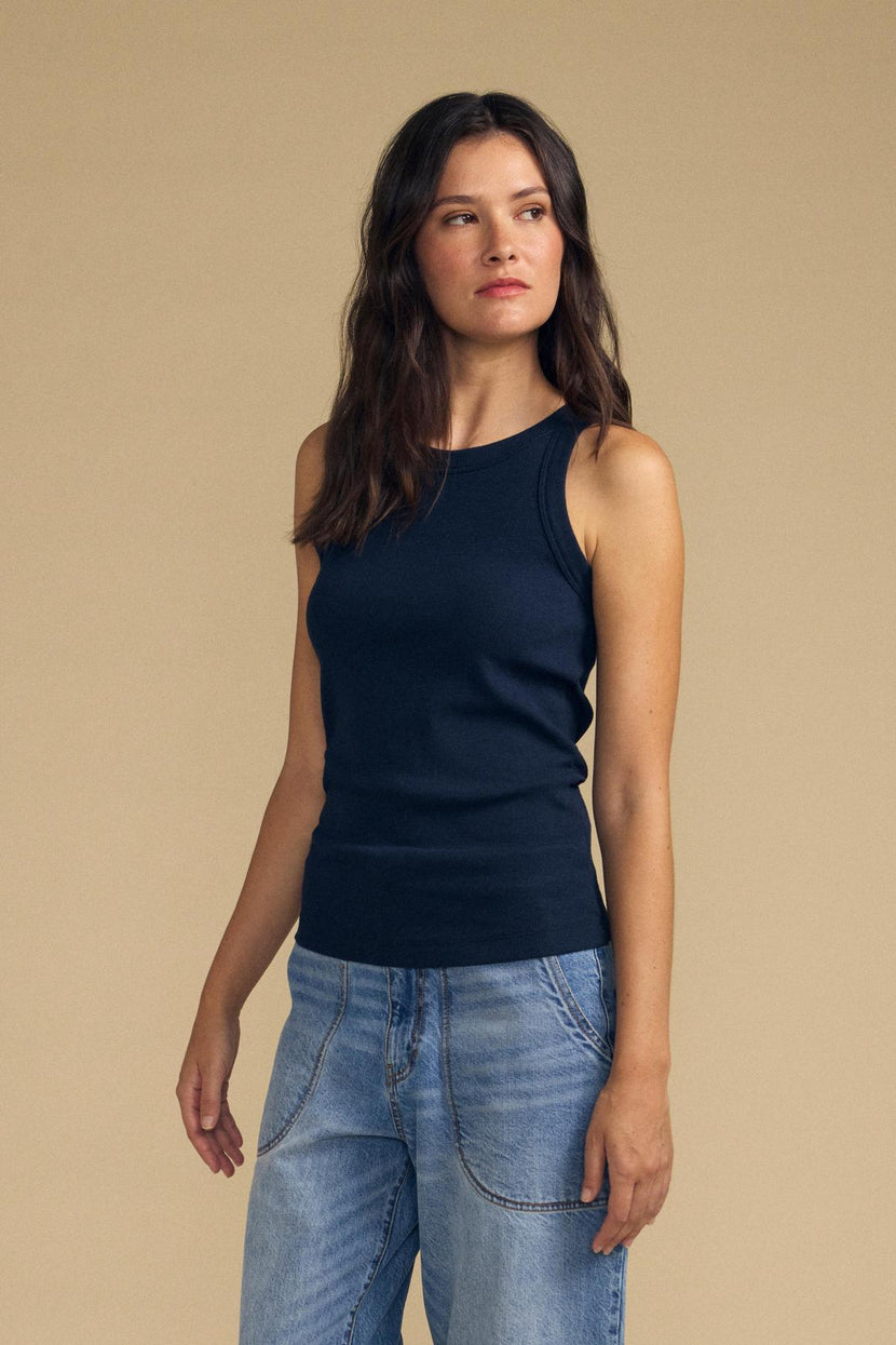 Wearing the CRUZ TANK TOP by Velvet by Jenny Graham, a woman with long brown hair styles the sleeveless, ribbed navy tank with light blue jeans against a plain beige background—a versatile look perfect for any capsule wardrobe.