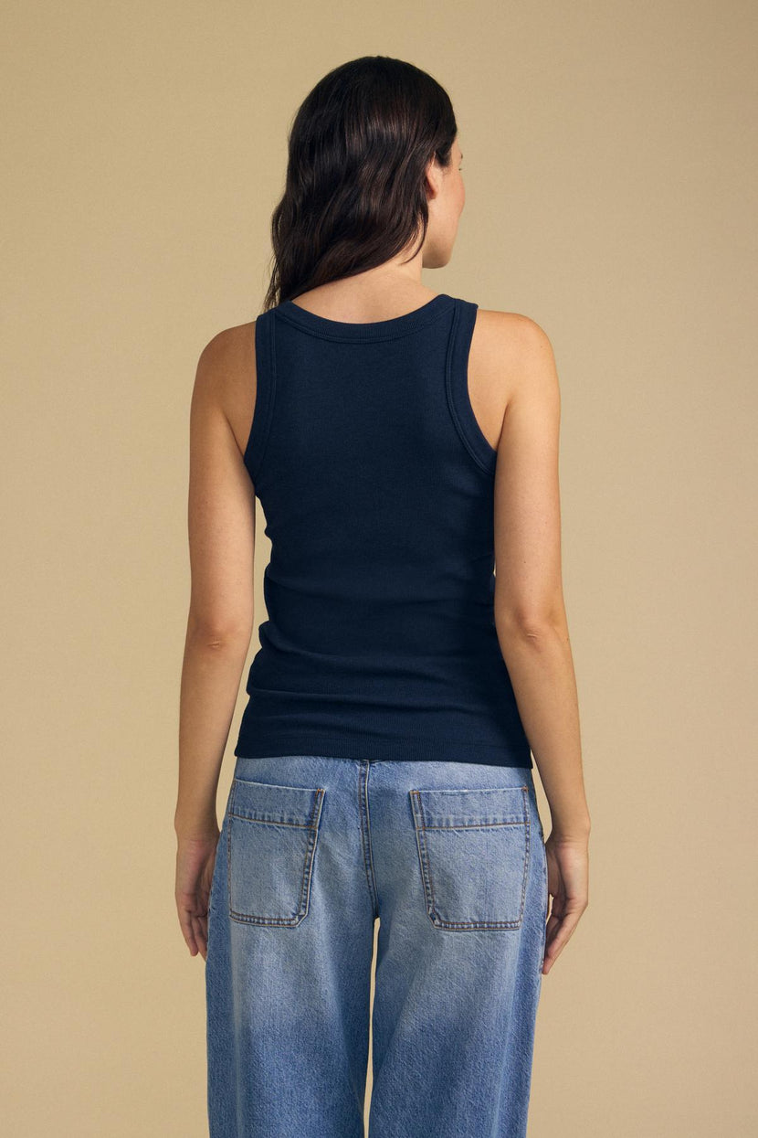 A woman stands facing away, wearing Velvet by Jenny Graham’s CRUZ TANK TOP in navy blue ribbed cotton-modal and light blue jeans against a beige background—an effortless staple for any capsule wardrobe.