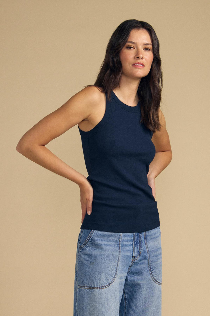 A person stands against a beige background, wearing the CRUZ TANK TOP in navy blue by Velvet by Jenny Graham from their capsule wardrobe and light blue jeans, hands resting on their hips.