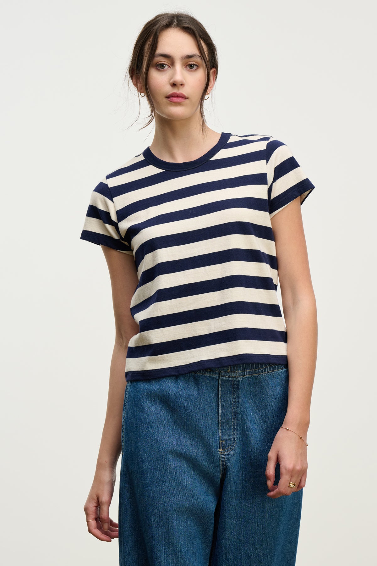   A woman stands against a plain background wearing the ANAIS TEE by Velvet by Graham & Spencer—a navy and white striped cotton jersey crewneck—paired with blue jeans. 