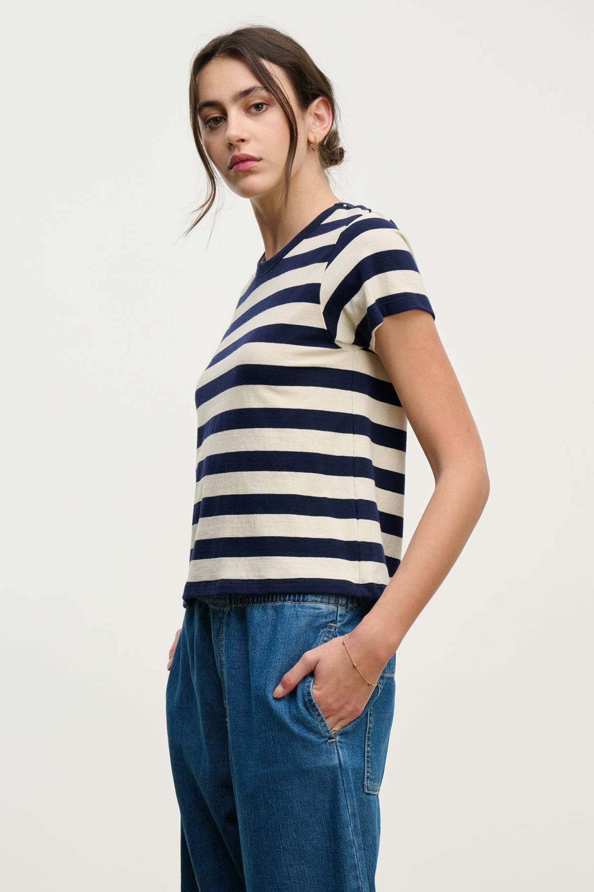   A woman wears the ANAIS TEE by Velvet by Graham & Spencer—a navy and white striped cotton jersey short sleeve crew neck—paired with blue jeans, standing with her hands in her pockets against a plain background. 