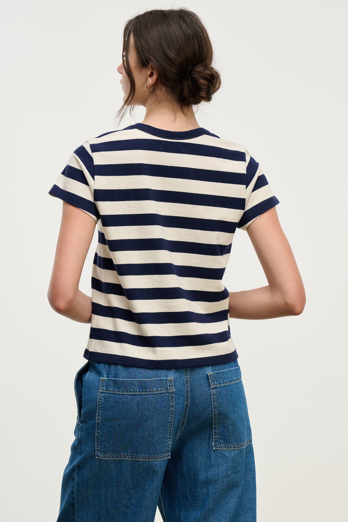 A person stands facing away, wearing the ANAIS TEE by Velvet by Graham & Spencer—a navy and white striped cotton jersey short sleeve crewneck—paired with blue jeans featuring large back pockets.-39725311328449