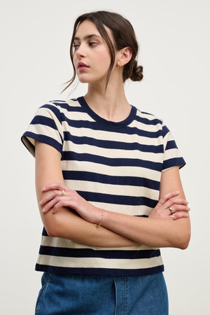 A woman in the ANAIS TEE by Velvet by Graham & Spencer, a soft cotton jersey with navy and white stripes and a classic crew neckline, stands with arms crossed against a plain background, wearing blue jeans.