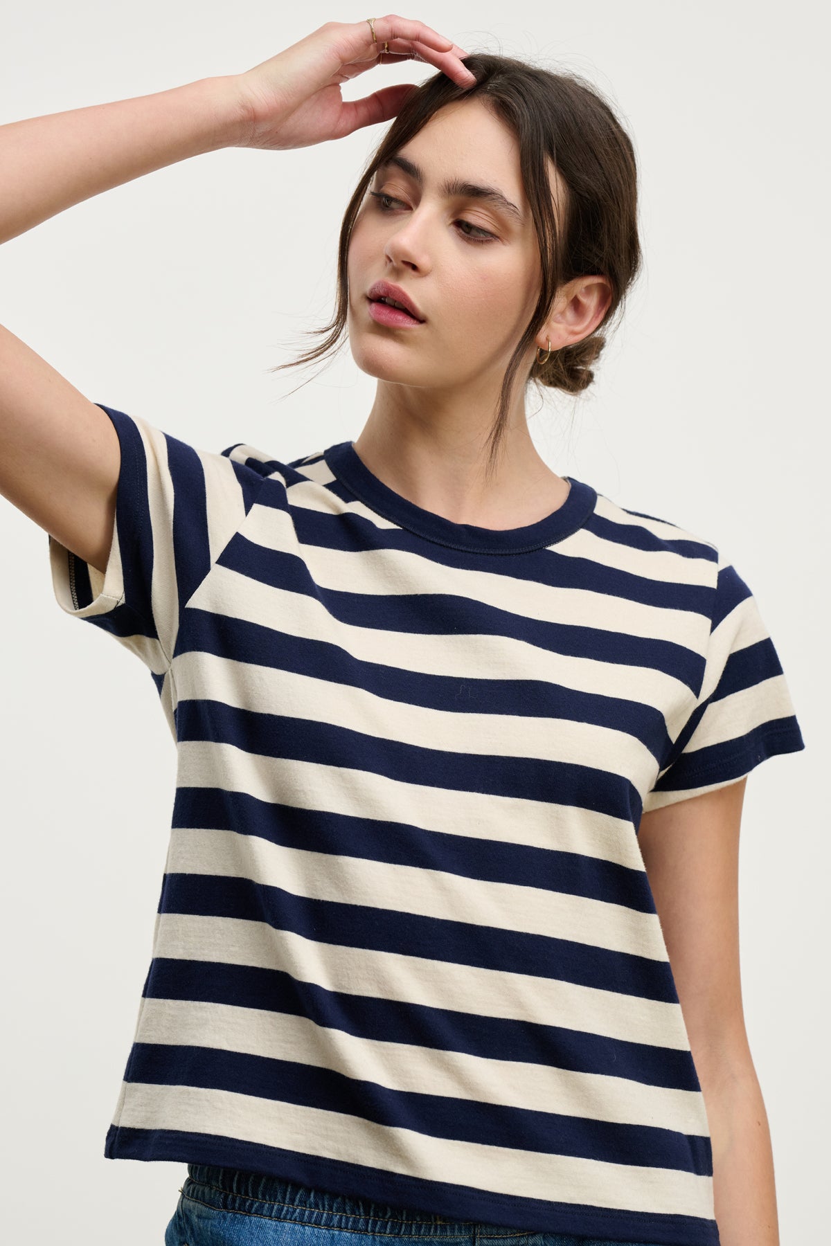   A person wears the ANAIS TEE by Velvet by Graham & Spencer—a navy and white striped cotton jersey short sleeve crewneck—standing against a plain background with one hand raised near their head. 