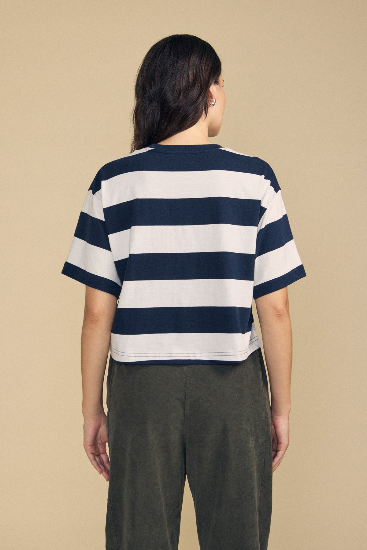 A person stands facing away against a plain beige background, wearing the FRYEE TEE by Velvet by Graham & Spencer—a navy and white striped cotton tee with a boxy fit—paired with dark green pants.