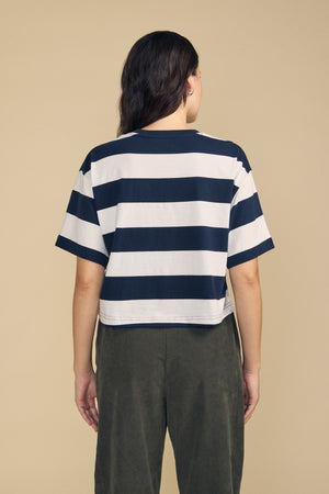 A person stands facing away against a plain beige background, wearing the FRYEE TEE by Velvet by Graham & Spencer—a navy and white striped cotton tee with a boxy fit—paired with dark green pants.