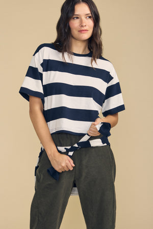 A woman stands against a beige background, holding the end of her FRYEE TEE by Velvet by Graham & Spencer—a black and white horizontally striped, boxy-fit cotton tee—paired with dark green pants.