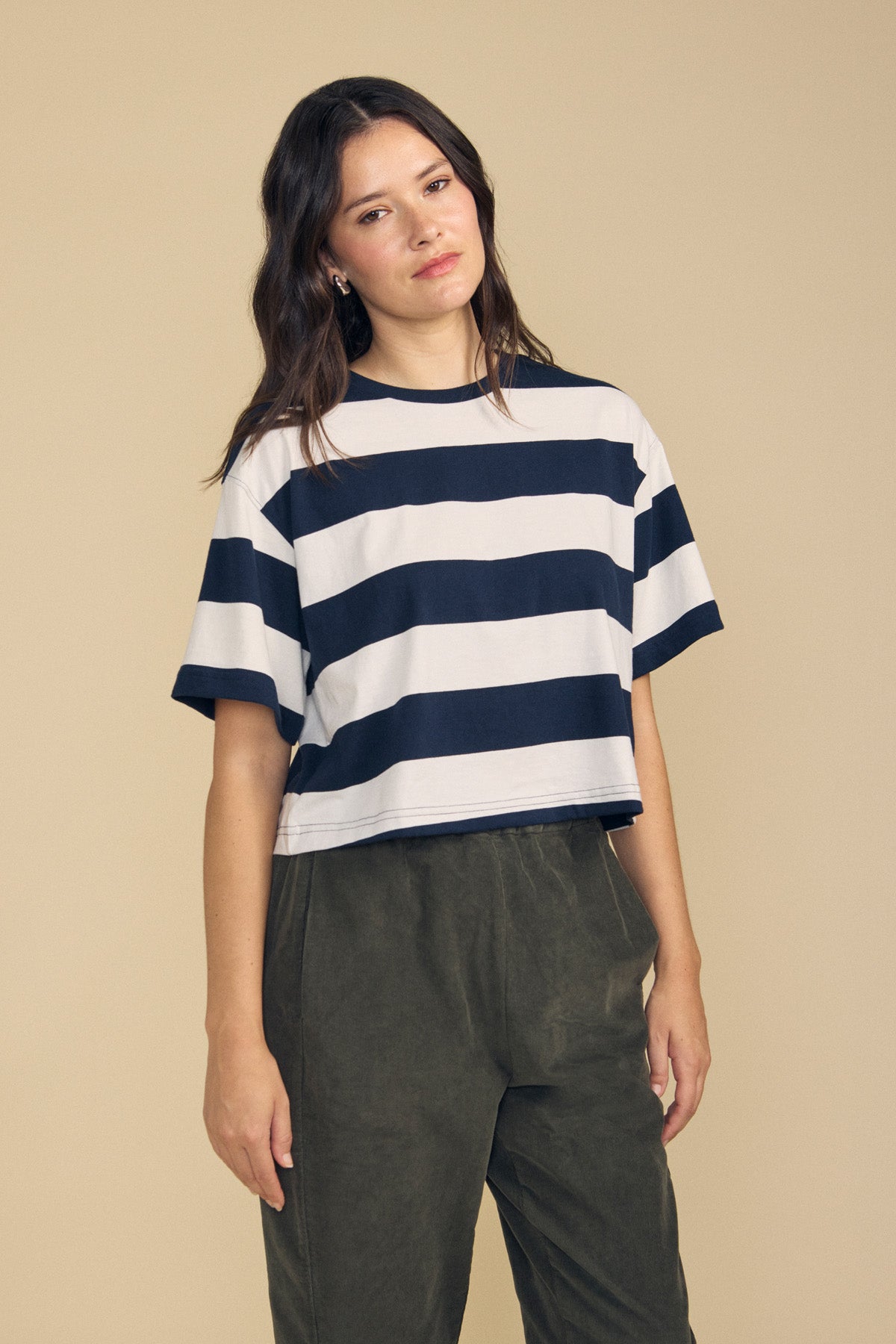 A woman stands against a plain beige background wearing the FRYEE TEE by Velvet by Graham & Spencer—a navy and white striped cotton tee with a boxy fit—paired with olive green pants.-41860431151297