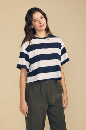 A woman stands against a plain beige background wearing the FRYEE TEE by Velvet by Graham & Spencer—a navy and white striped cotton tee with a boxy fit—paired with olive green pants.
