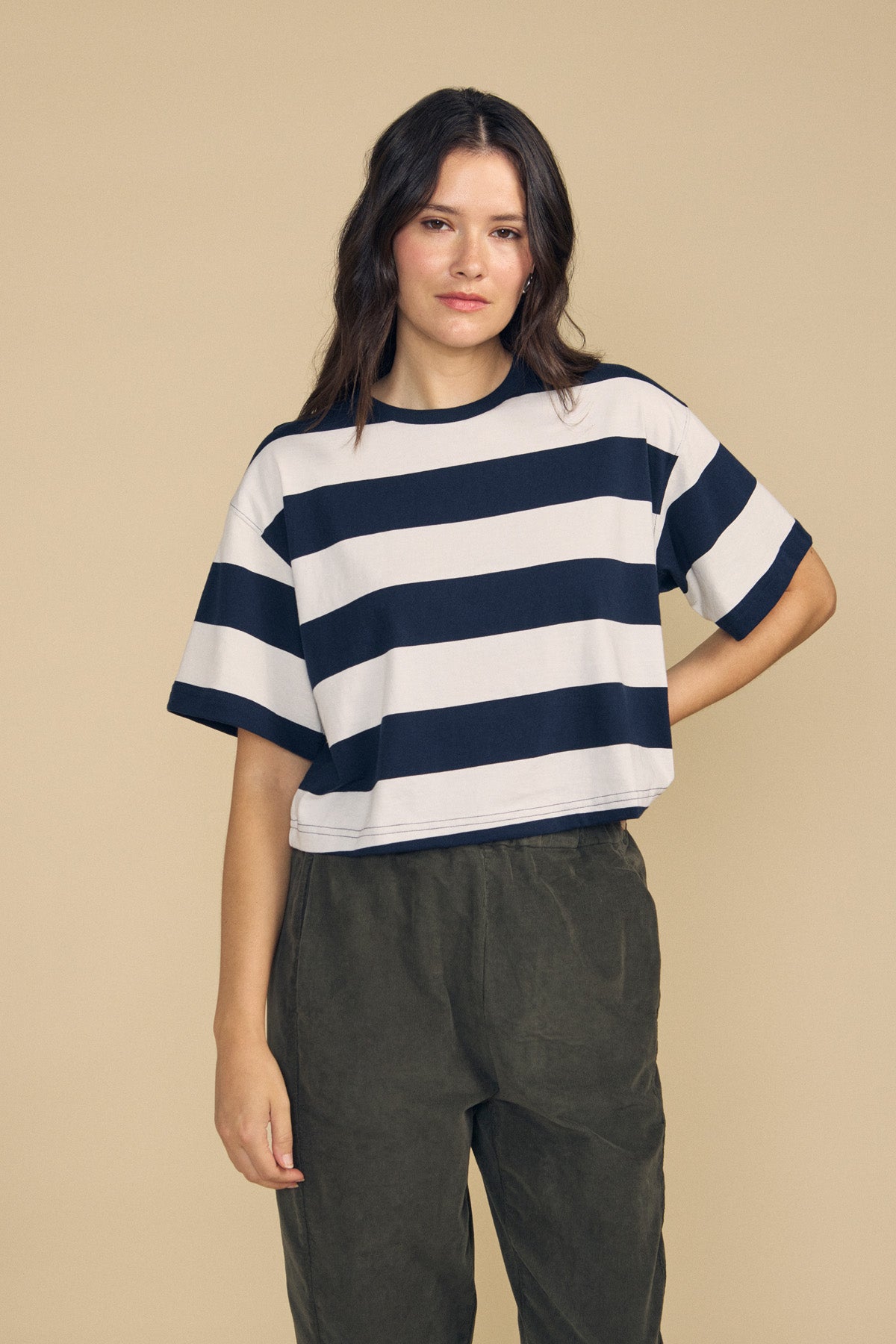 A person stands against a beige background wearing the FRYEE TEE by Velvet by Graham & Spencer—a loose, boxy-fit short-sleeve shirt featuring wide black and white horizontal stripes—paired with dark green pants.-41860431118529