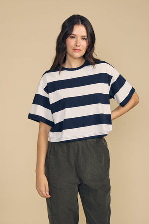 A person stands against a beige background wearing the FRYEE TEE by Velvet by Graham & Spencer—a loose, boxy-fit short-sleeve shirt featuring wide black and white horizontal stripes—paired with dark green pants.