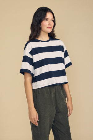 A woman stands in front of a beige background wearing the FRYEE TEE by Velvet by Graham & Spencer—a cropped-length, black-and-white striped cotton jersey top with a boxy, relaxed fit—paired with olive green pants.