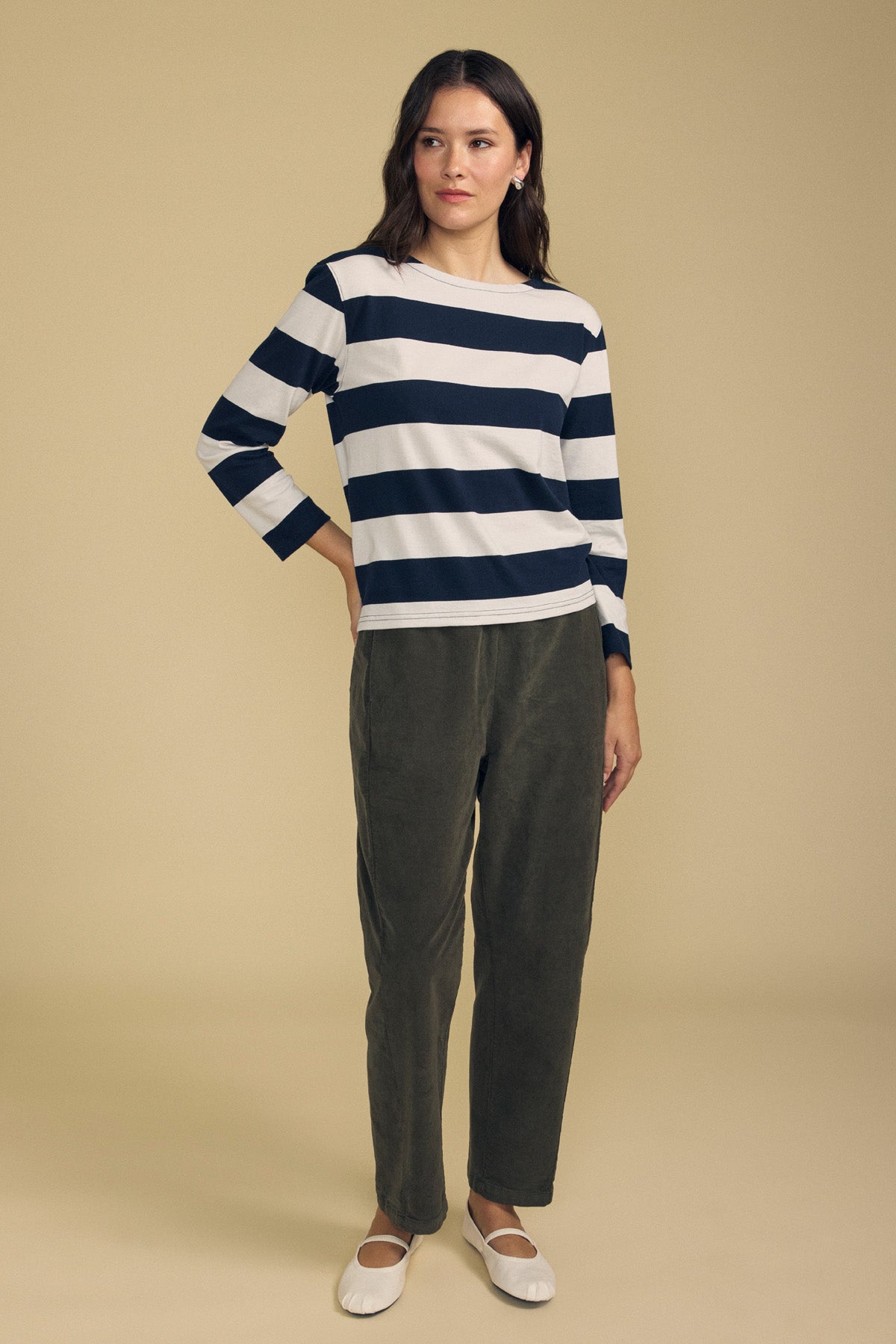 Woman standing against a beige background, wearing a navy and white striped long-sleeve shirt, Velvet by Graham & Spencer LANEY CORDUROY PANT in olive green with a pull-on waist, and white ballet flats.