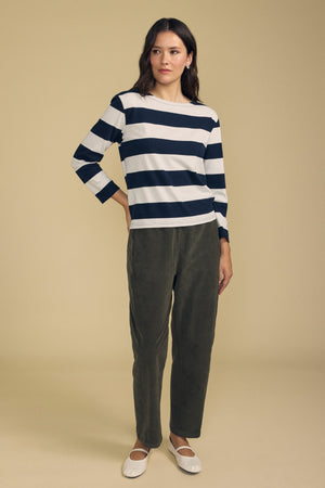 Woman standing against a beige background, wearing a navy and white striped long-sleeve shirt, Velvet by Graham & Spencer LANEY CORDUROY PANT in olive green with a pull-on waist, and white ballet flats.