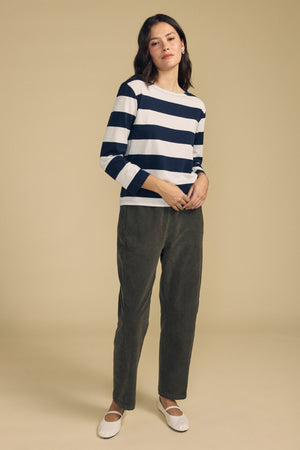 Person standing against a beige background, wearing the GABRIELA TEE by Velvet by Graham & Spencer—a relaxed fit, long sleeve navy and white striped cotton jersey top—paired with olive green pants and white shoes.