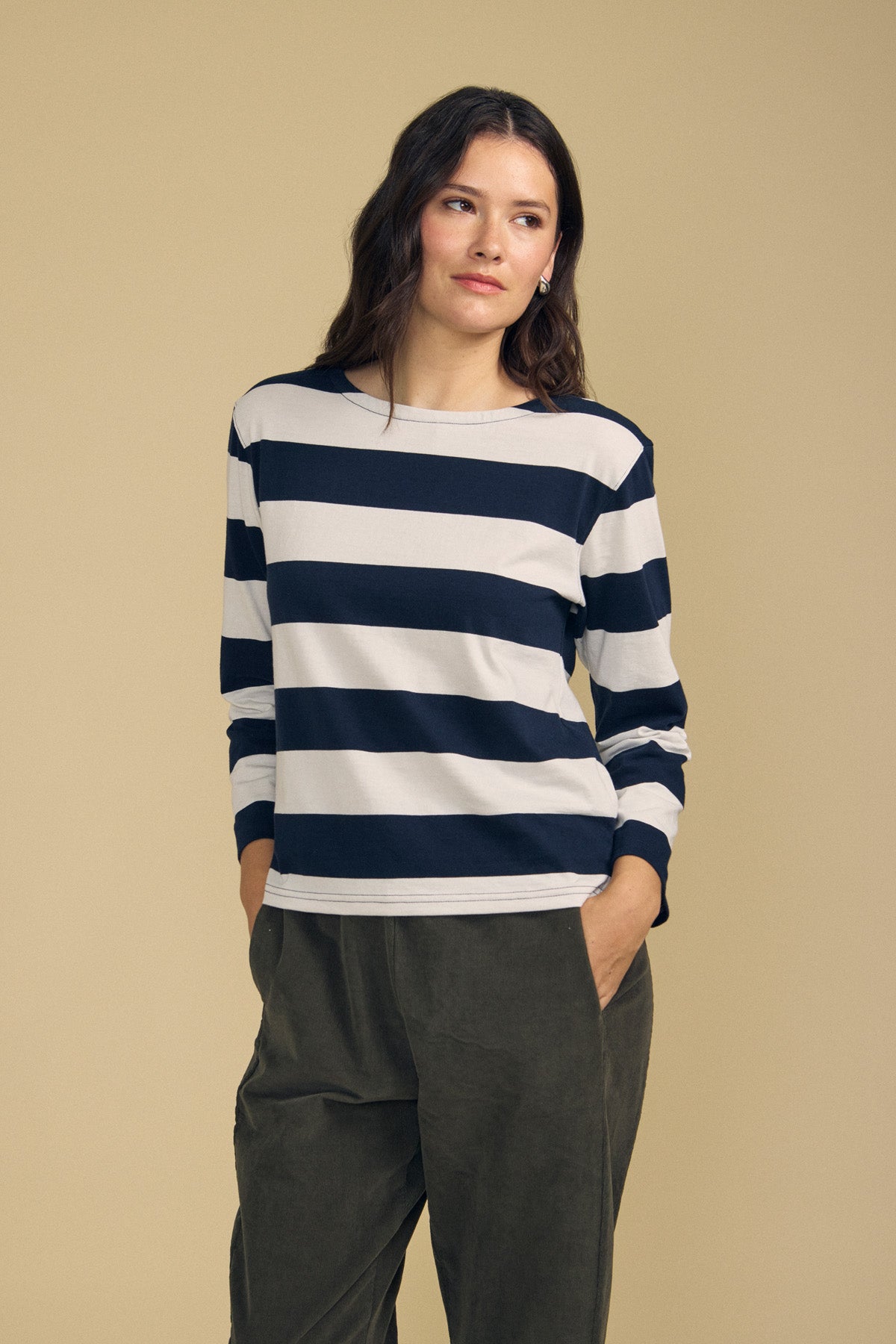 A woman stands against a beige background wearing the Velvet by Graham & Spencer GABRIELA TEE—a navy and white long sleeve striped cotton jersey top—paired with olive green pants, her hands in her pockets for a relaxed fit look.