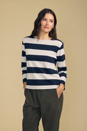 A woman stands against a beige background wearing the Velvet by Graham & Spencer GABRIELA TEE—a navy and white long sleeve striped cotton jersey top—paired with olive green pants, her hands in her pockets for a relaxed fit look.