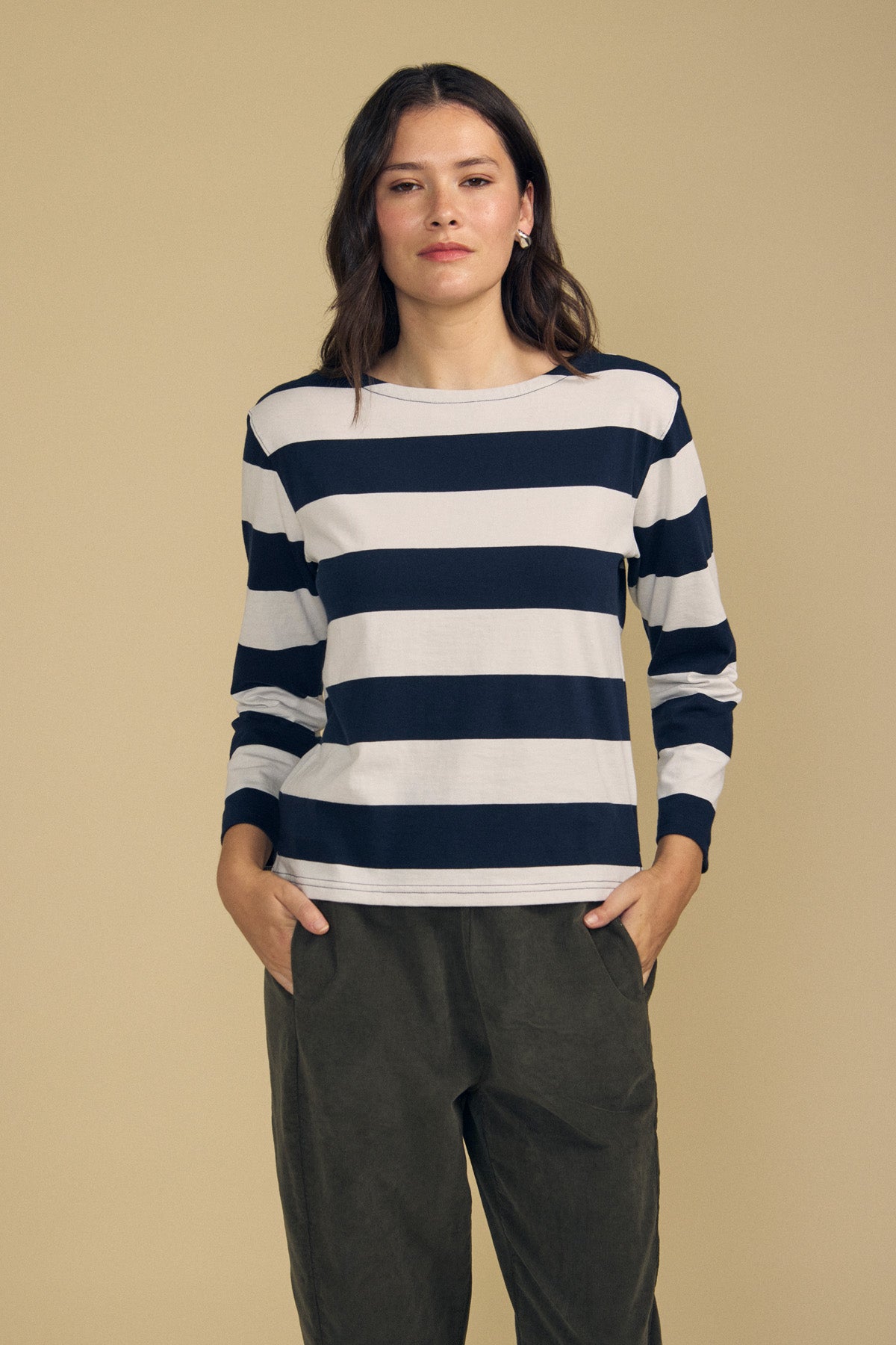 A woman stands against a beige background, wearing the GABRIELA TEE by Velvet by Graham & Spencer—a relaxed fit, navy and white striped cotton jersey long-sleeve top—paired with olive green pants and her hands in her pockets.