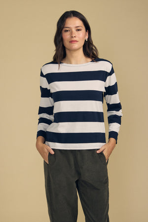 A woman stands against a beige background, wearing the GABRIELA TEE by Velvet by Graham & Spencer—a relaxed fit, navy and white striped cotton jersey long-sleeve top—paired with olive green pants and her hands in her pockets.